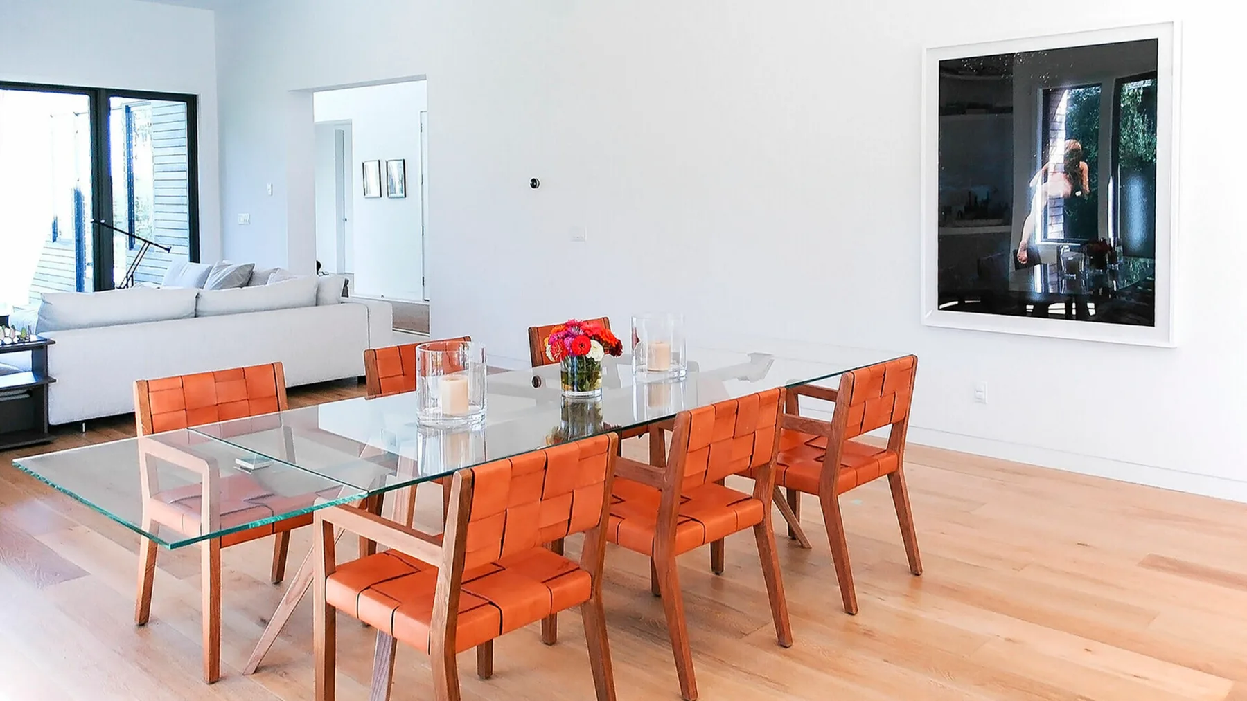 Un comedor luminoso con mesa de vidrio y asientos naranjas da a una sala de estar.