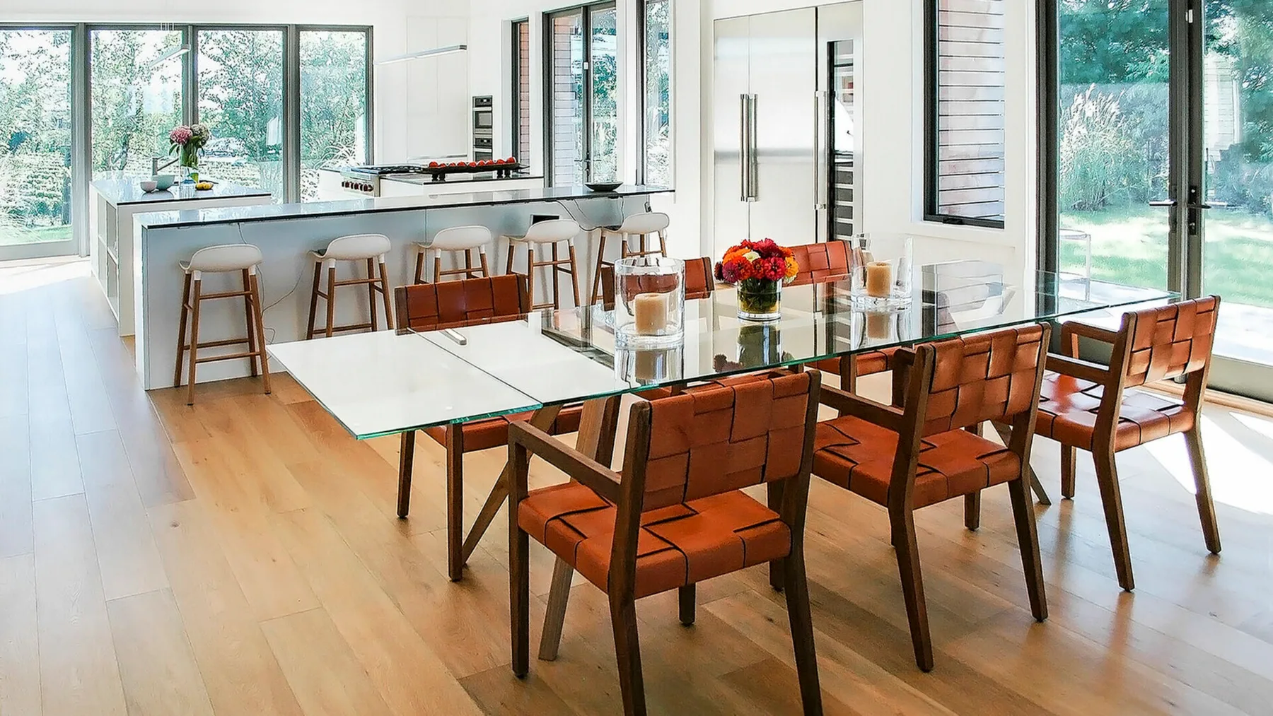 Un comedor luminoso con una mesa de vidrio y asientos marrones junto a grandes ventanales.