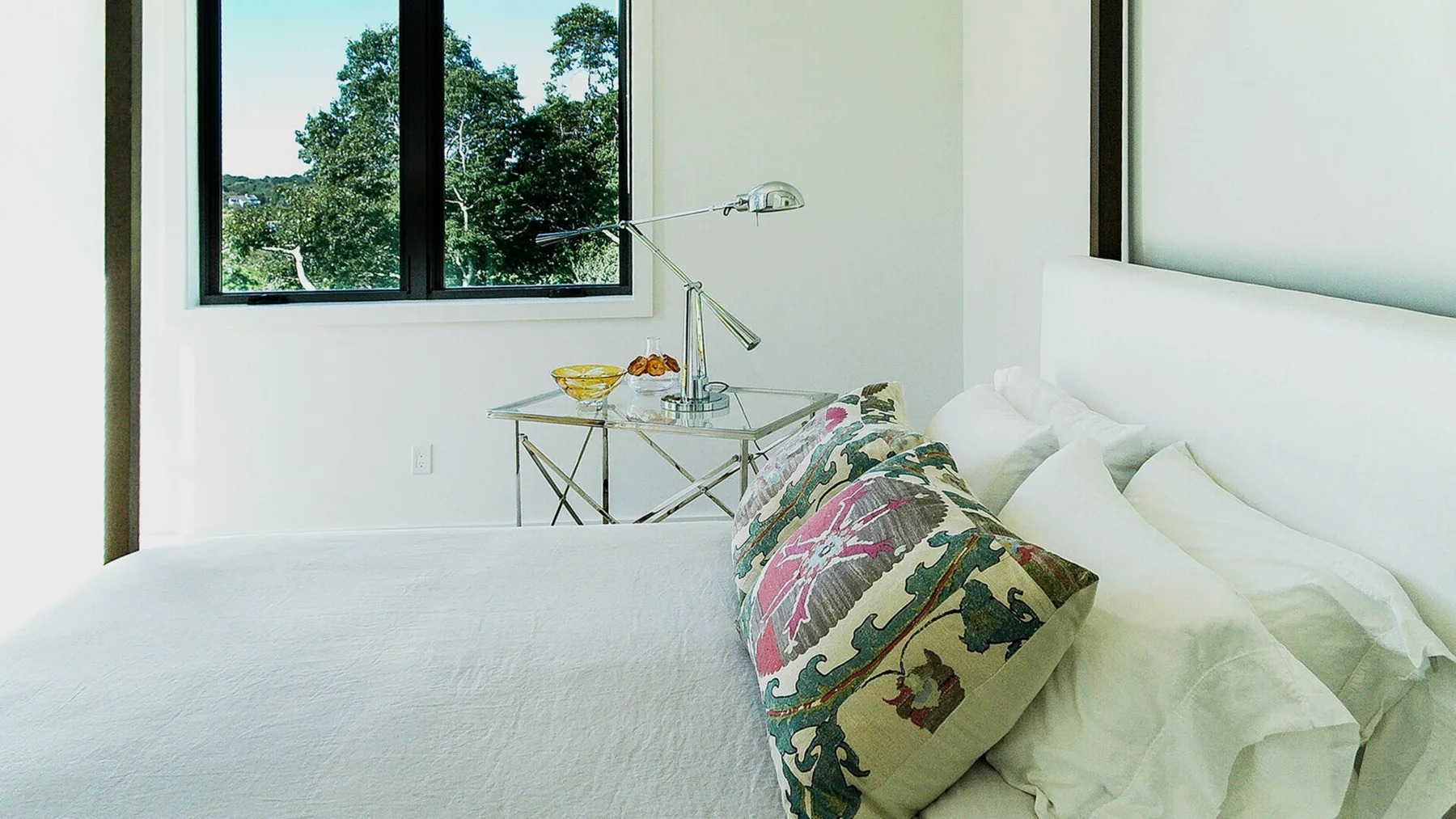 Un dormitorio luminoso con ropa de cama blanca y un cojín estampado junto a una ventana.