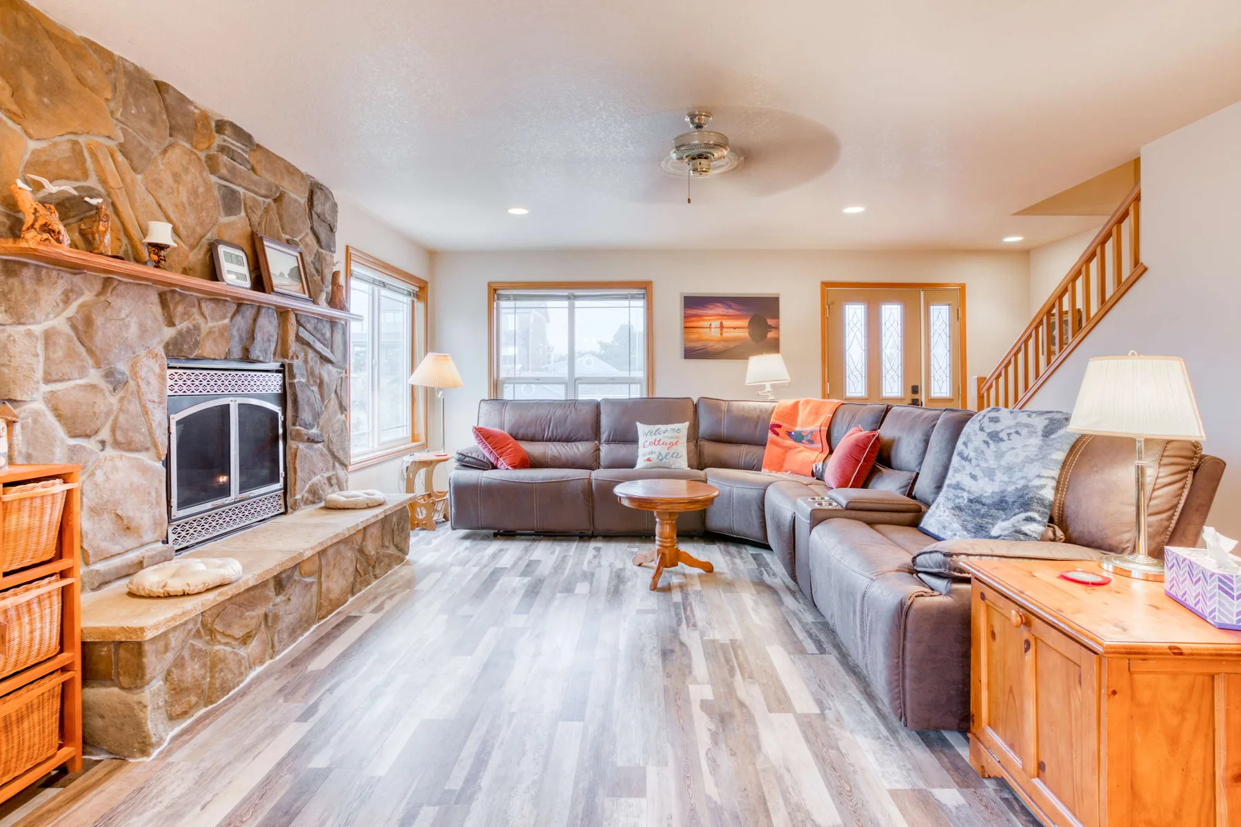 A living room with sectional seating, a stone fireplace, and a wooden staircase.