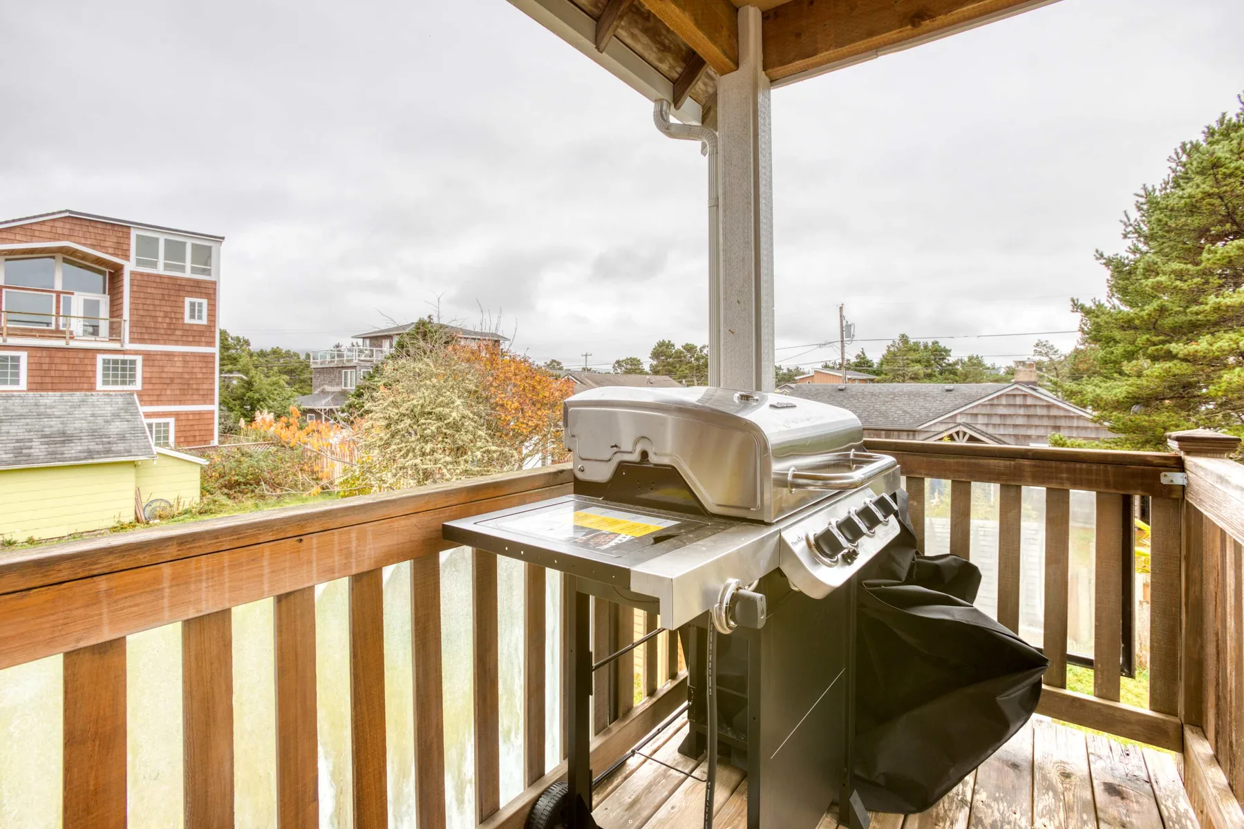 A wooden deck with a covered grill and railing overlooking nearby houses and trees.