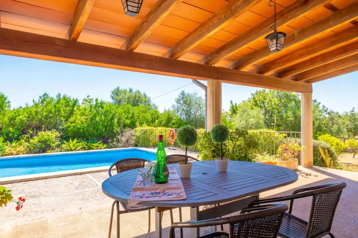 Una zona de comedor en un patio cubierto da a una piscina y un jardín frondoso.