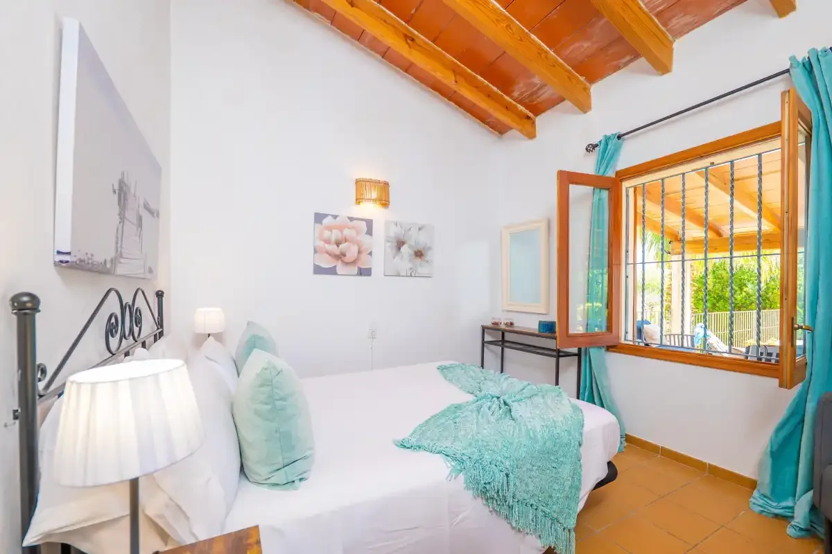 Un dormitorio luminoso con ropa de cama blanca, detalles turquesa y una ventana grande abierta.