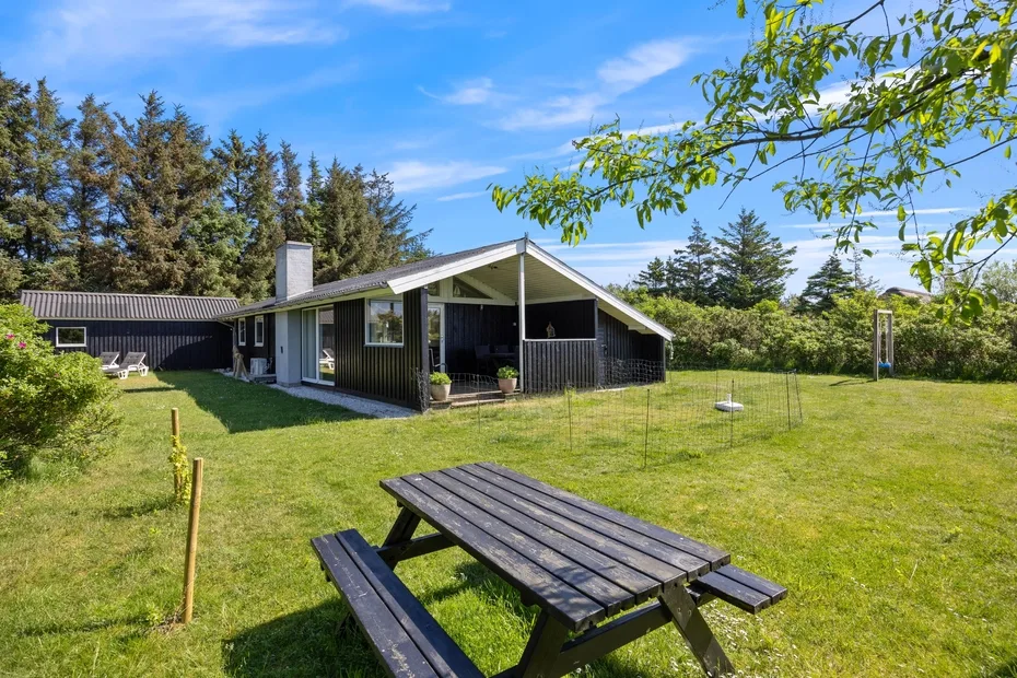 Una casa de revestimiento oscuro y una mesa de picnic están en un patio soleado con césped.