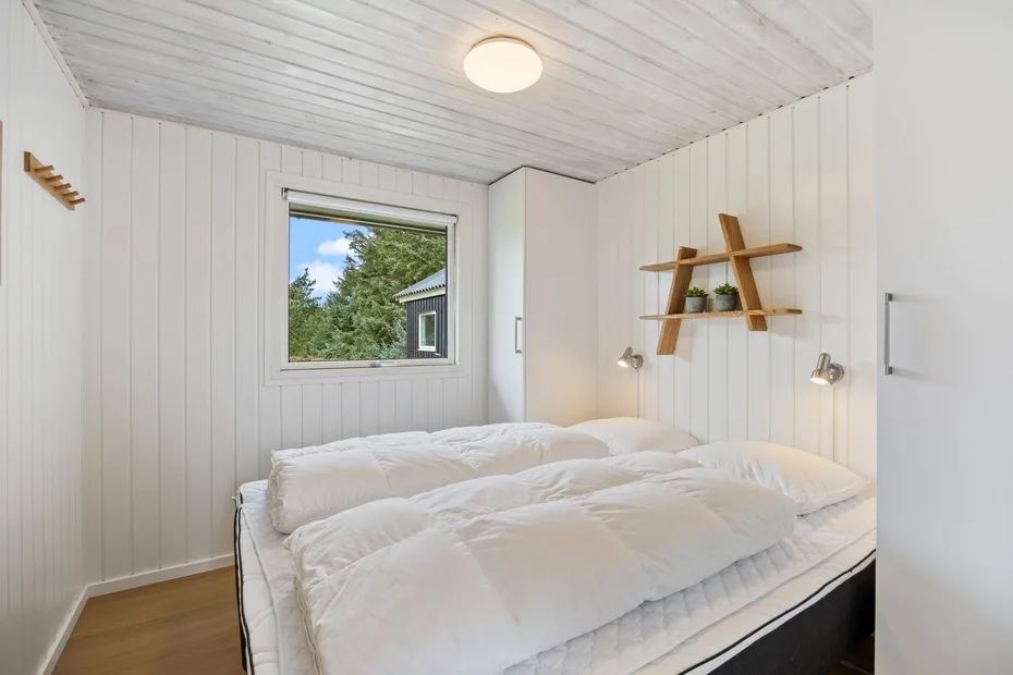 Un dormitorio luminoso con ropa de cama blanca y una ventana con vista a los árboles.