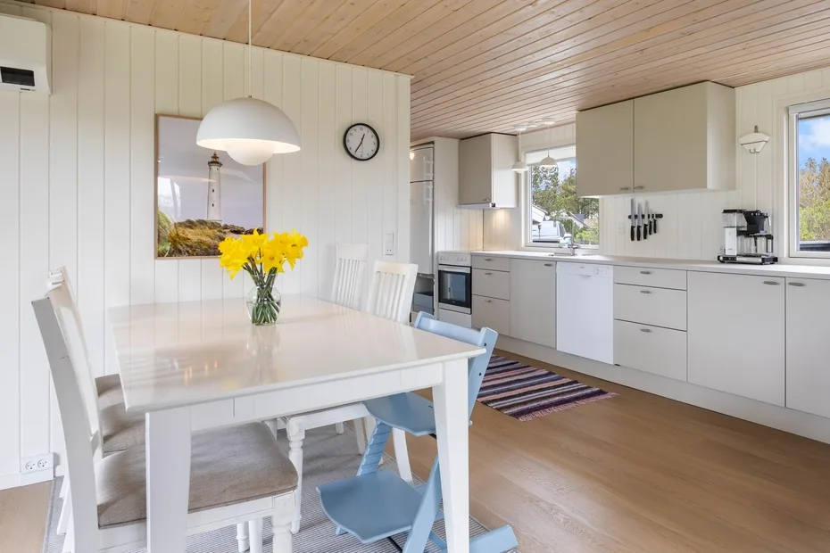 Un comedor luminoso se abre a una cocina blanca con una mesa y flores de colores.