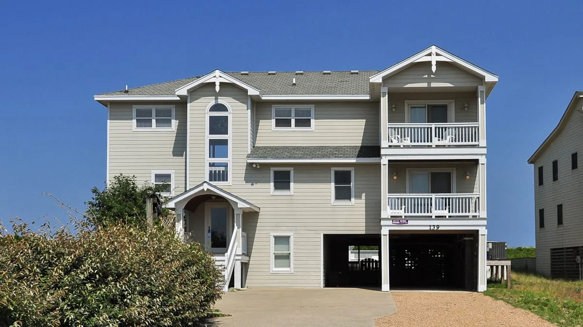 A large light-colored house with balconies stands beside a paved driveway.