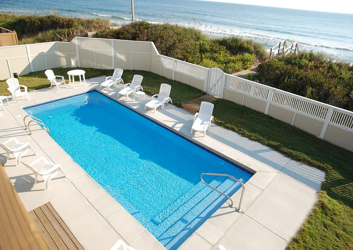 An outdoor swimming pool is surrounded by lounge chairs, with the ocean visible beyond the fence.