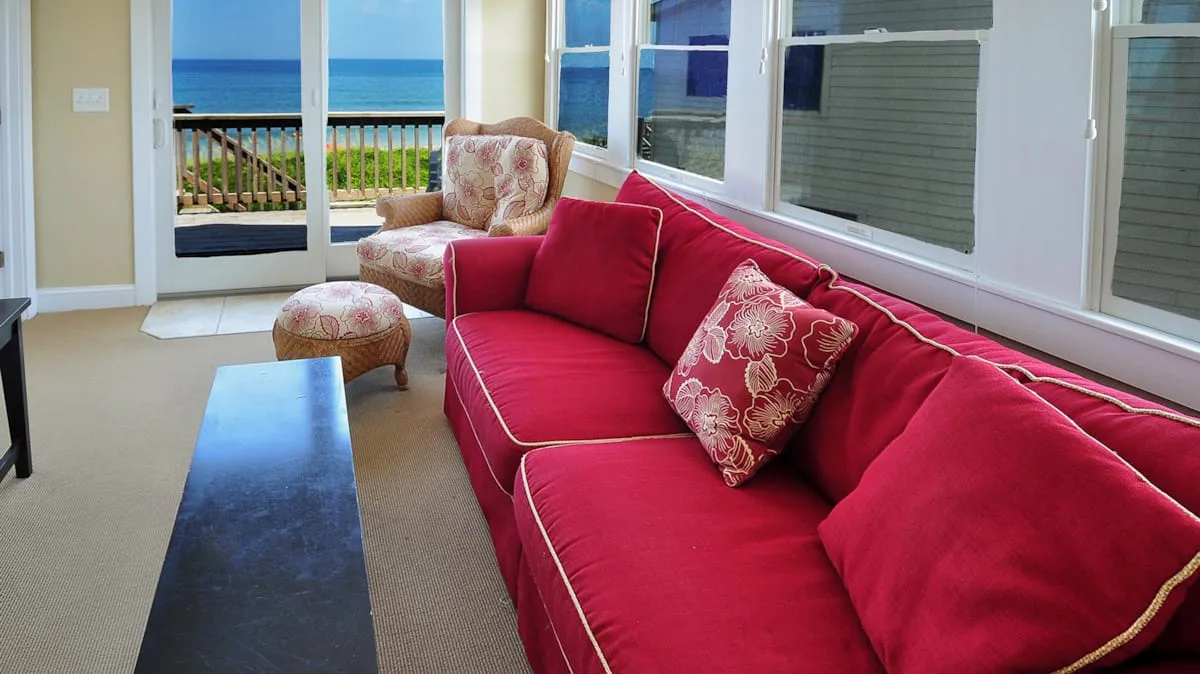 A bright sitting room has red seating beside a large window overlooking the ocean.