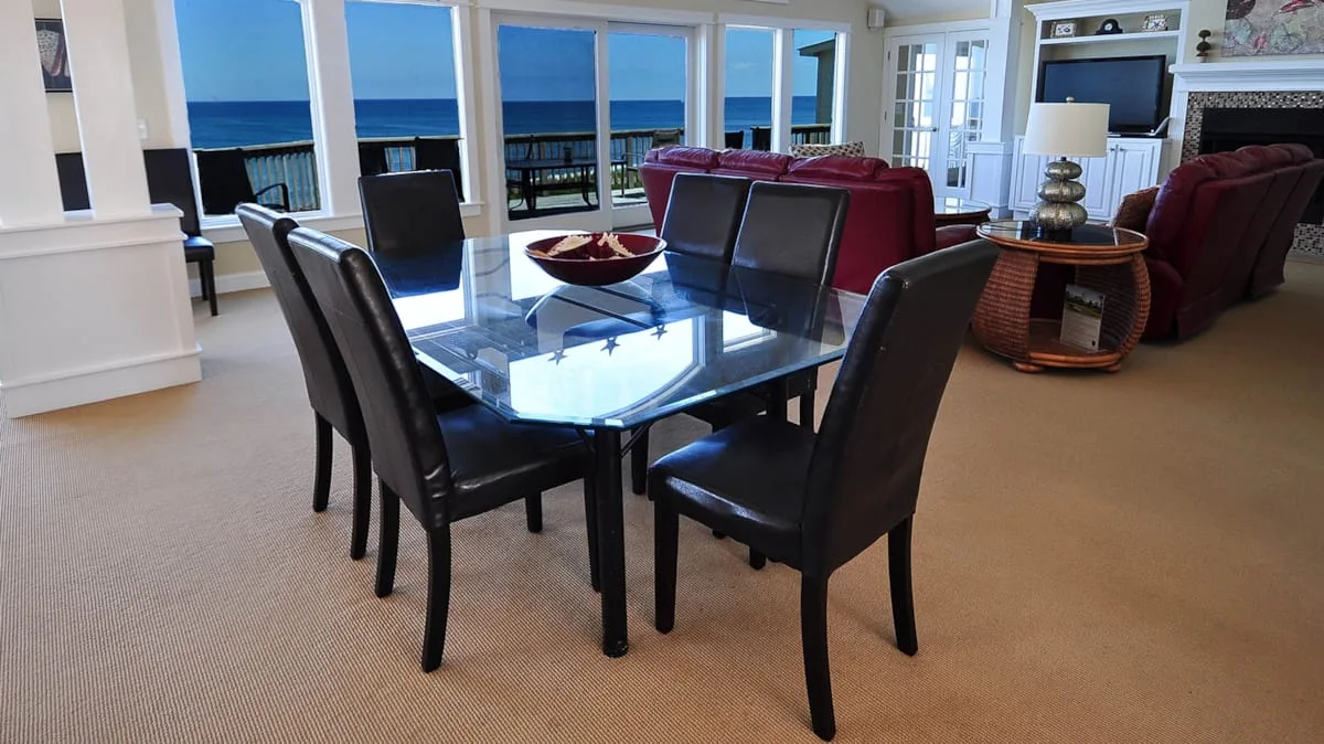 A dining area with a glass table and seating beside large windows overlooking the ocean.
