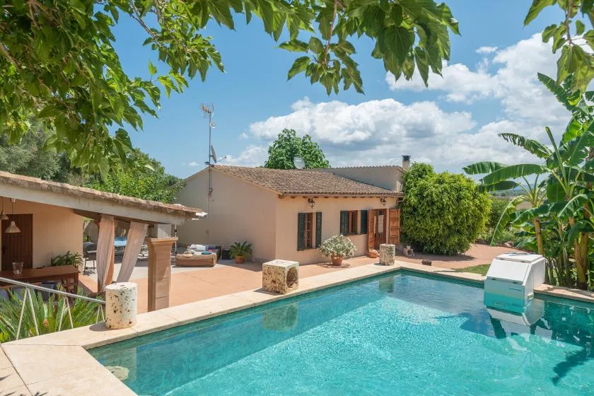 Una piscina junto a una casa de una planta y un patio sombreado.