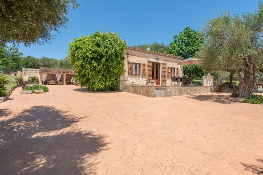 Una casa de piedra se encuentra junto a un amplio patio pavimentado rodeado de árboles.