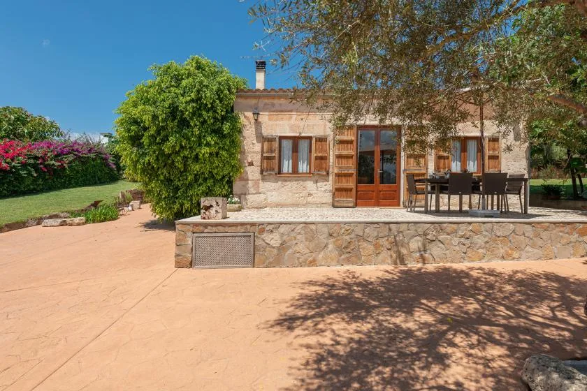 Una casa de piedra da a un patio pavimentado con asientos al aire libre y árboles.