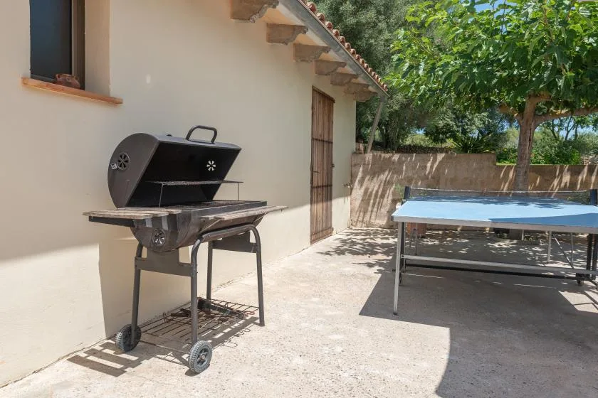 Un patio exterior soleado tiene una parrilla junto a una mesa de ping-pong.