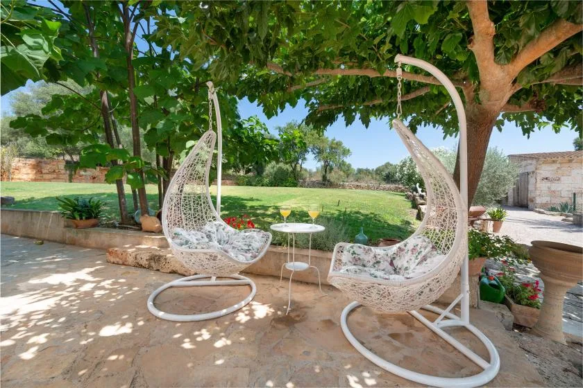 Un patio al aire libre con asientos colgantes bajo árboles frondosos, frente a un jardín verde.