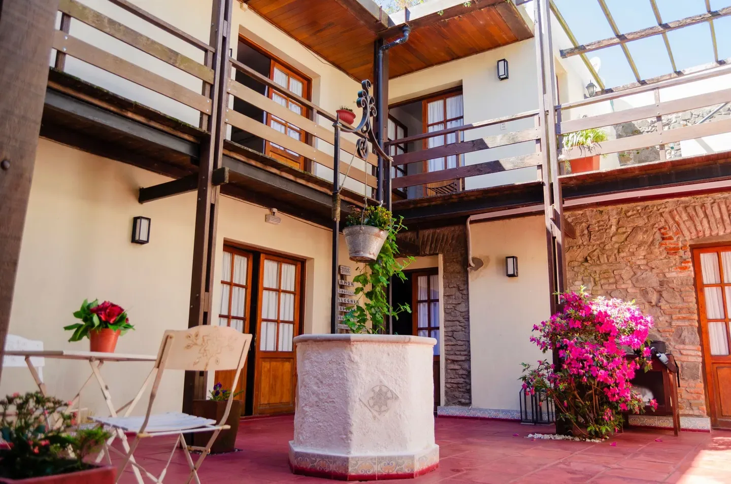 Patio con un pozo de piedra, balcones de madera, una mesa pequeña y enredaderas en flor.