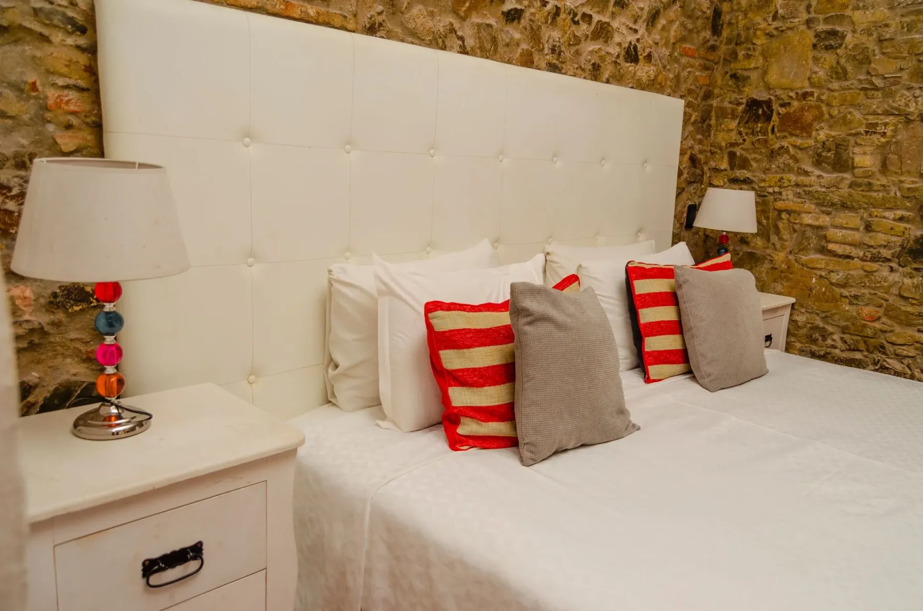 Cama junto a una pared de piedra con cabecero tapizado en blanco.