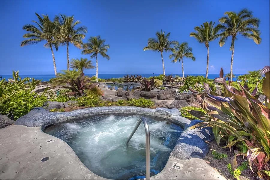 Un spa exterior burbujeante está rodeado de plantas tropicales, con palmeras y el océano al fondo.