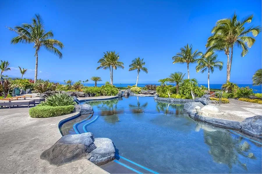 Una piscina de resort serpentea entre jardines tropicales, palmeras y vistas al mar.