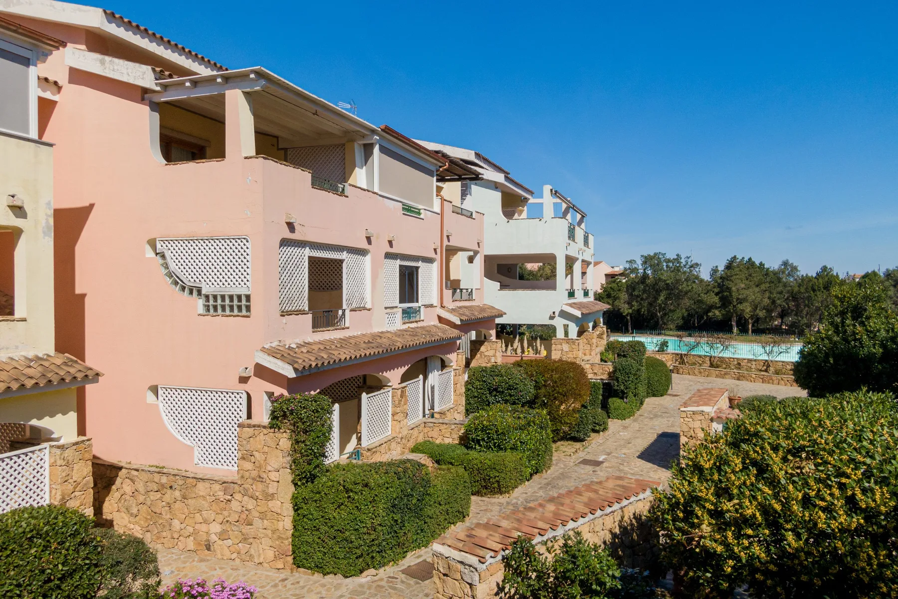 A pink stucco building stands beside a paved garden path and green shrubs.