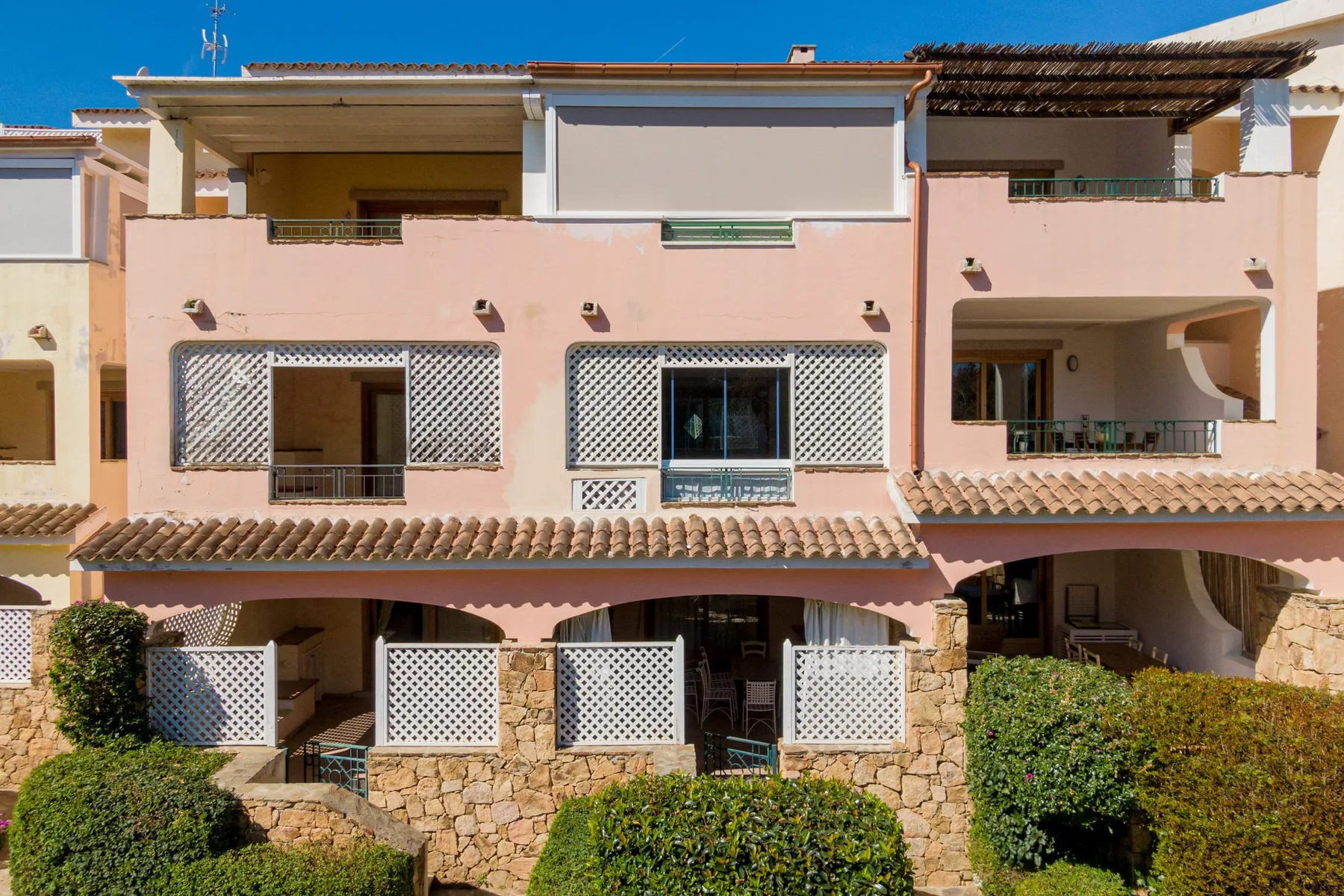 A pink multi-story residential building with balconies and trimmed shrubs in front.