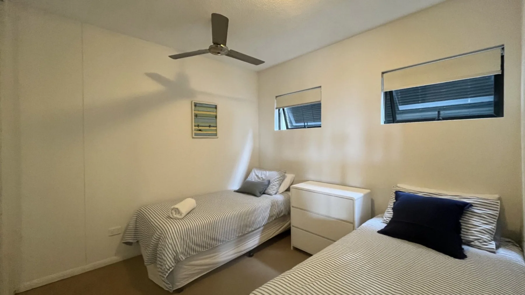 A bedroom with two beds, a bedside cabinet, and high windows.