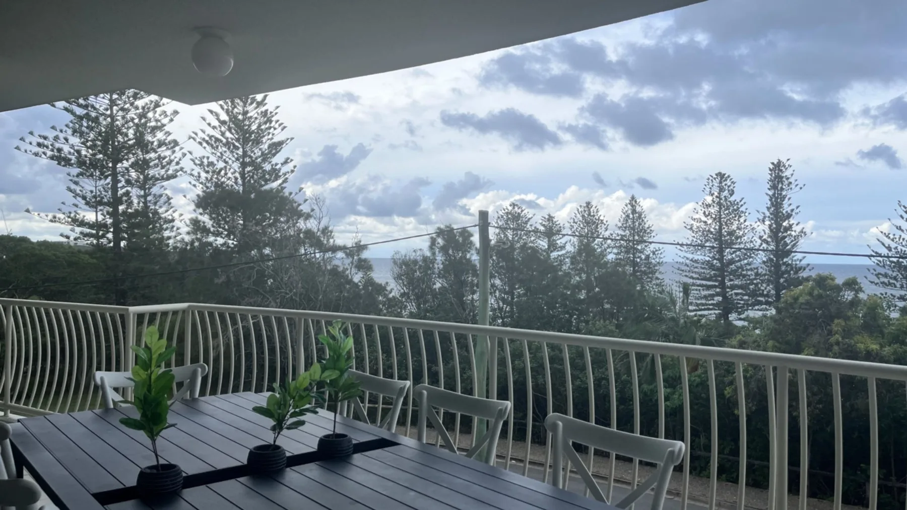 A covered balcony overlooks trees and greenery, with outdoor seating and potted plants.