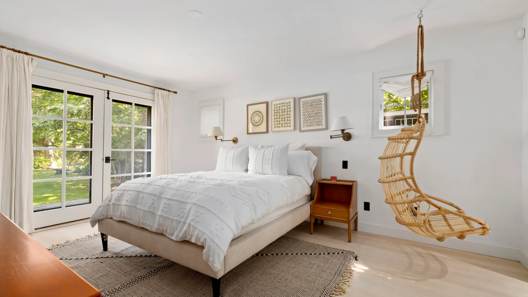 Un dormitorio luminoso con una cama blanca y un asiento colgante de mimbre.