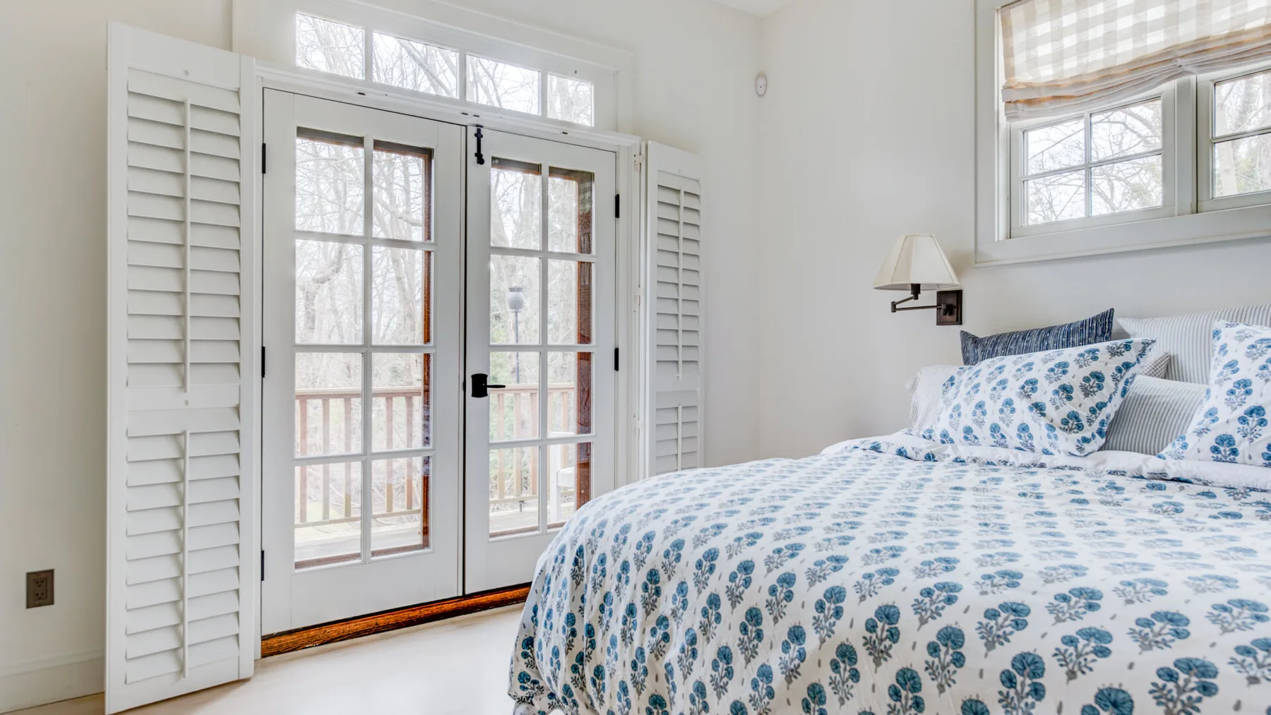 Un dormitorio luminoso con una cama junto a puertas acristaladas y postigos blancos.