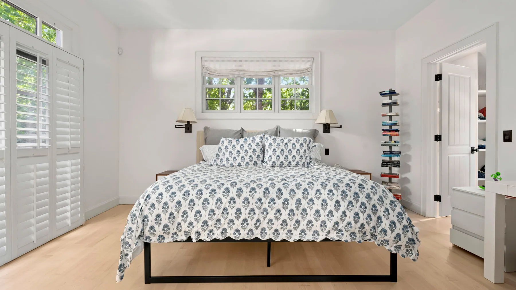 Un dormitorio luminoso con una cama bajo una ventana y contraventanas blancas.