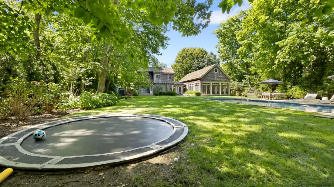Un patio ajardinado con trampolín circular, césped y piscina.