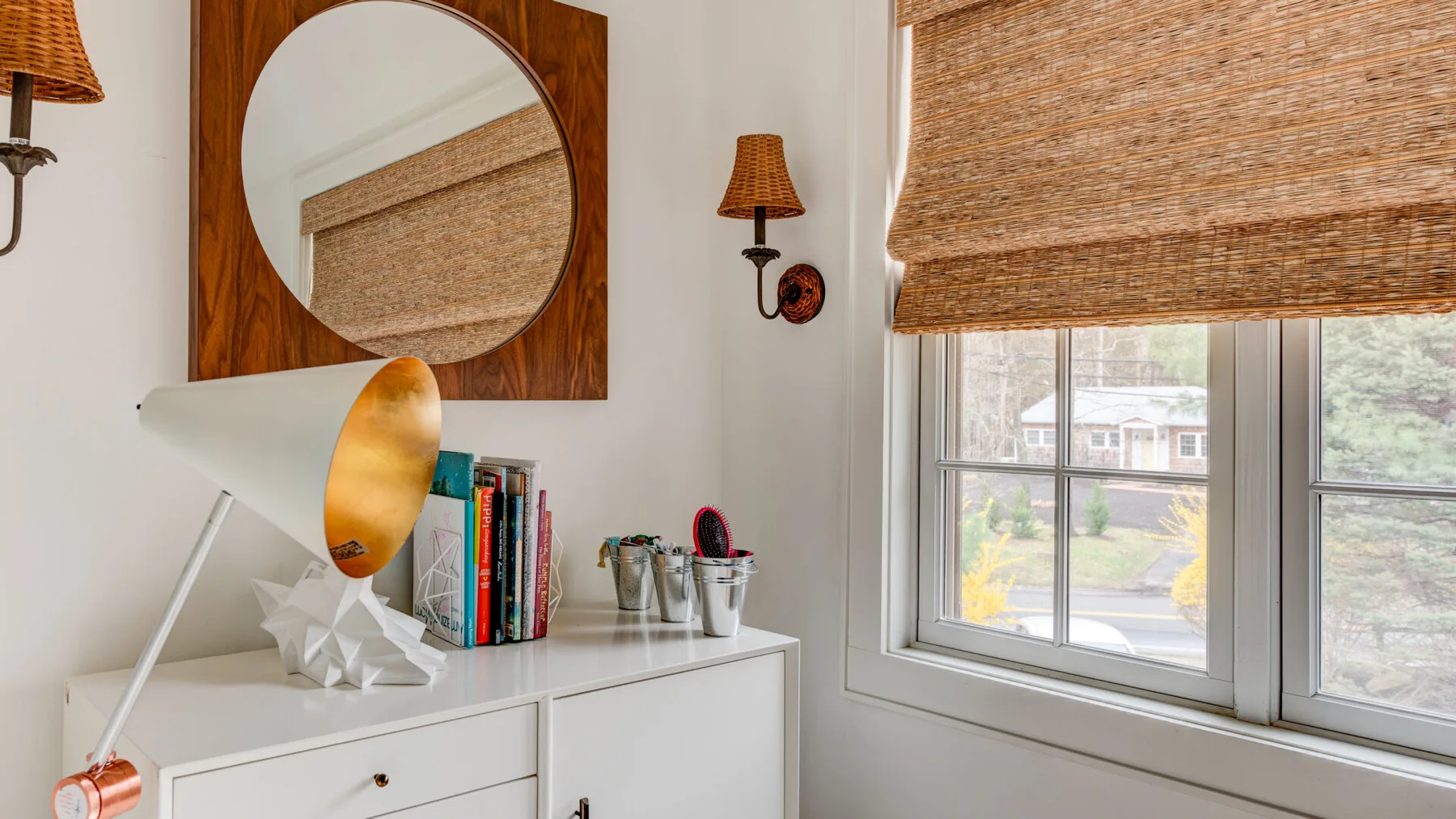 Un rincón luminoso con escritorio blanco, libros y una ventana con persianas tejidas.