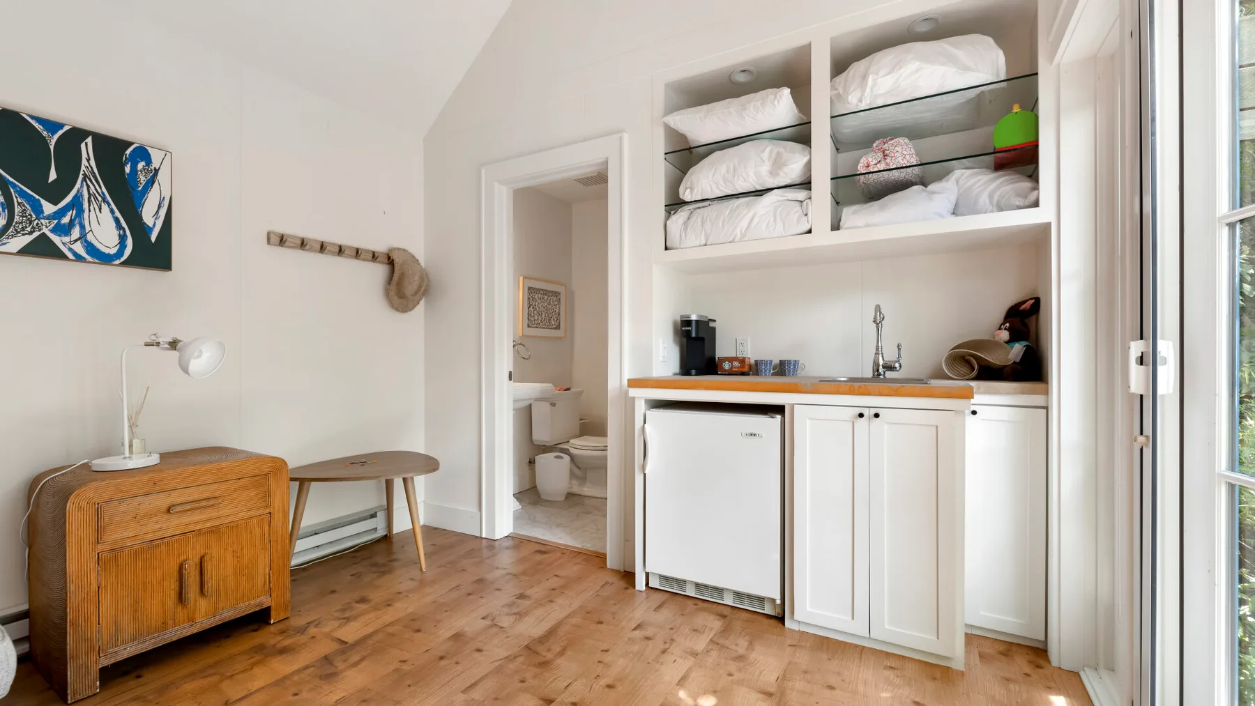 Una pequeña cocina con gabinetes blancos y estantes abiertos está junto a una habitación luminosa.