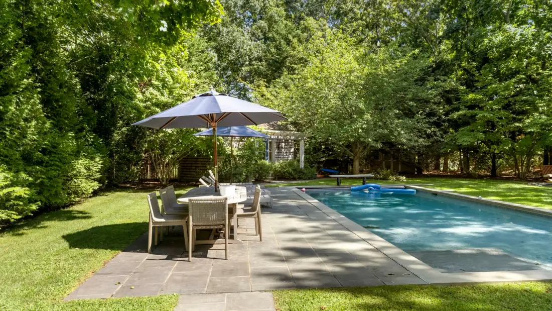 Una piscina al aire libre junto a un patio pavimentado con mesa y sombrilla.