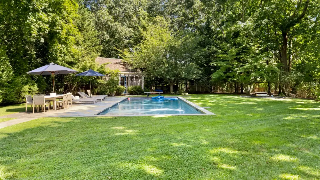 Una piscina y un patio están junto a un amplio césped rodeado de árboles.