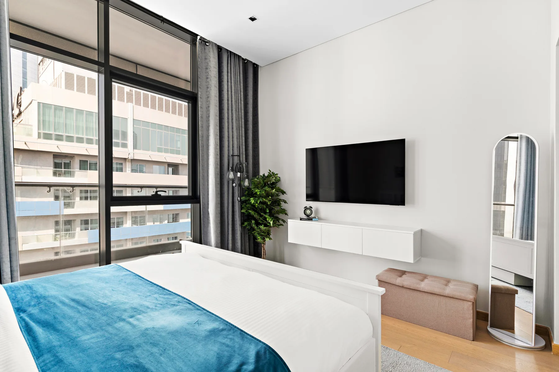 A bright bedroom with a large bed, wall-mounted television, and floor-to-ceiling windows.