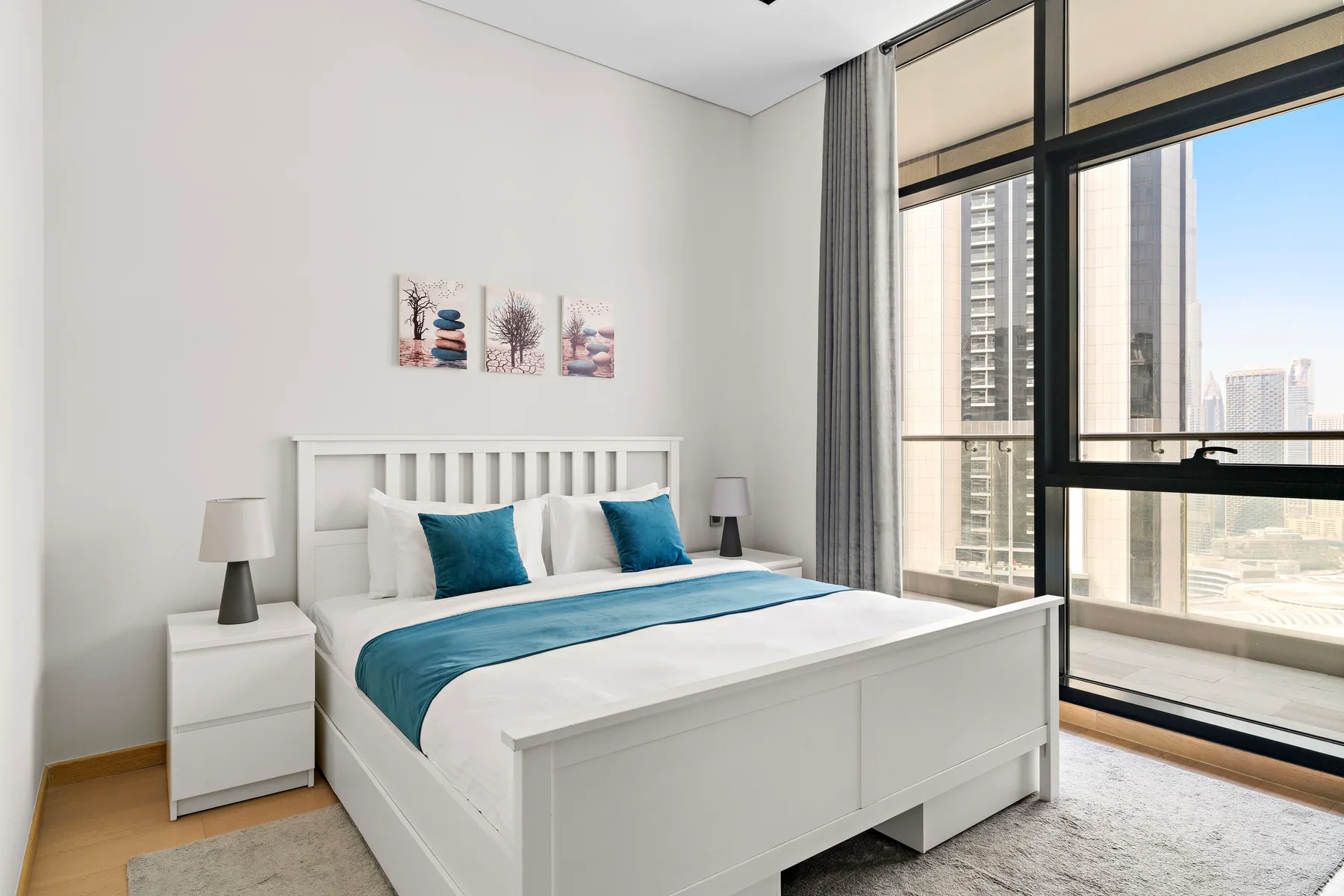 A bright bedroom with a white bed and large windows overlooking nearby buildings.