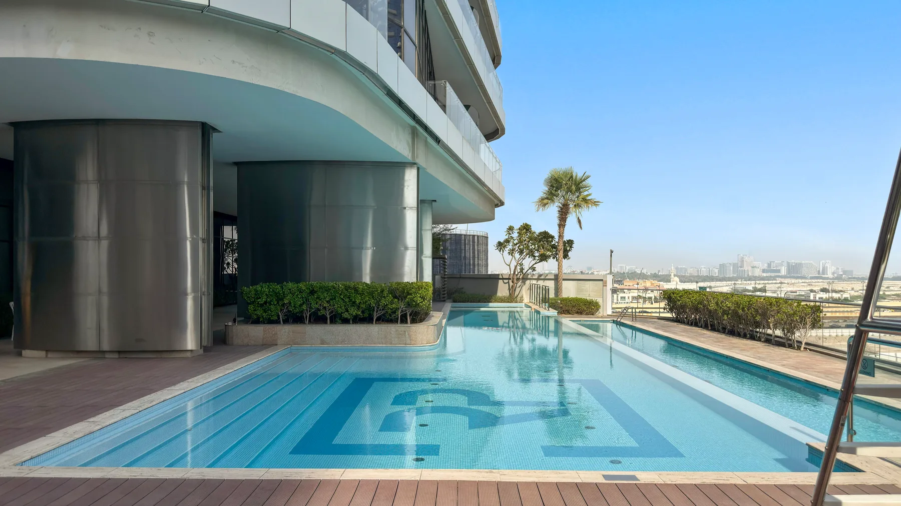 An outdoor swimming pool beside a modern building, with palm trees and a distant city view.