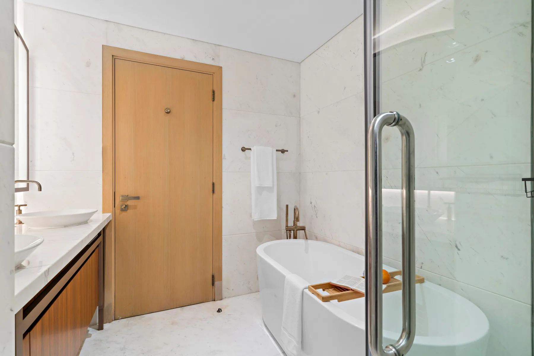 A bright bathroom with a bathtub, glass shower screen, and wood-tone door.