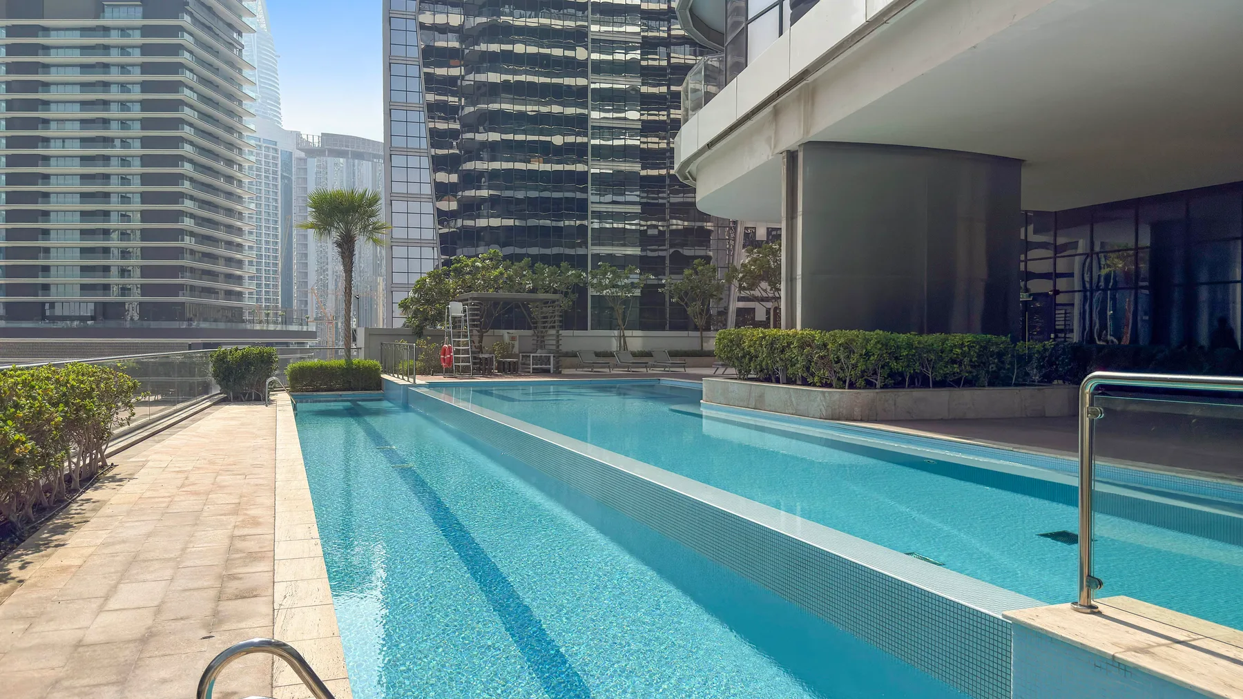 An outdoor swimming pool sits beside a paved walkway and modern buildings.