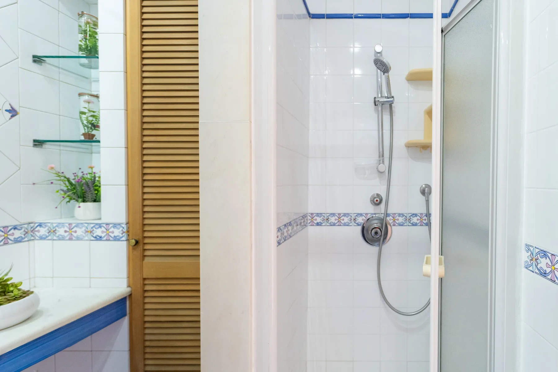 Un baño alicatado con ducha de vidrio, ducha de mano y estantes en la pared.