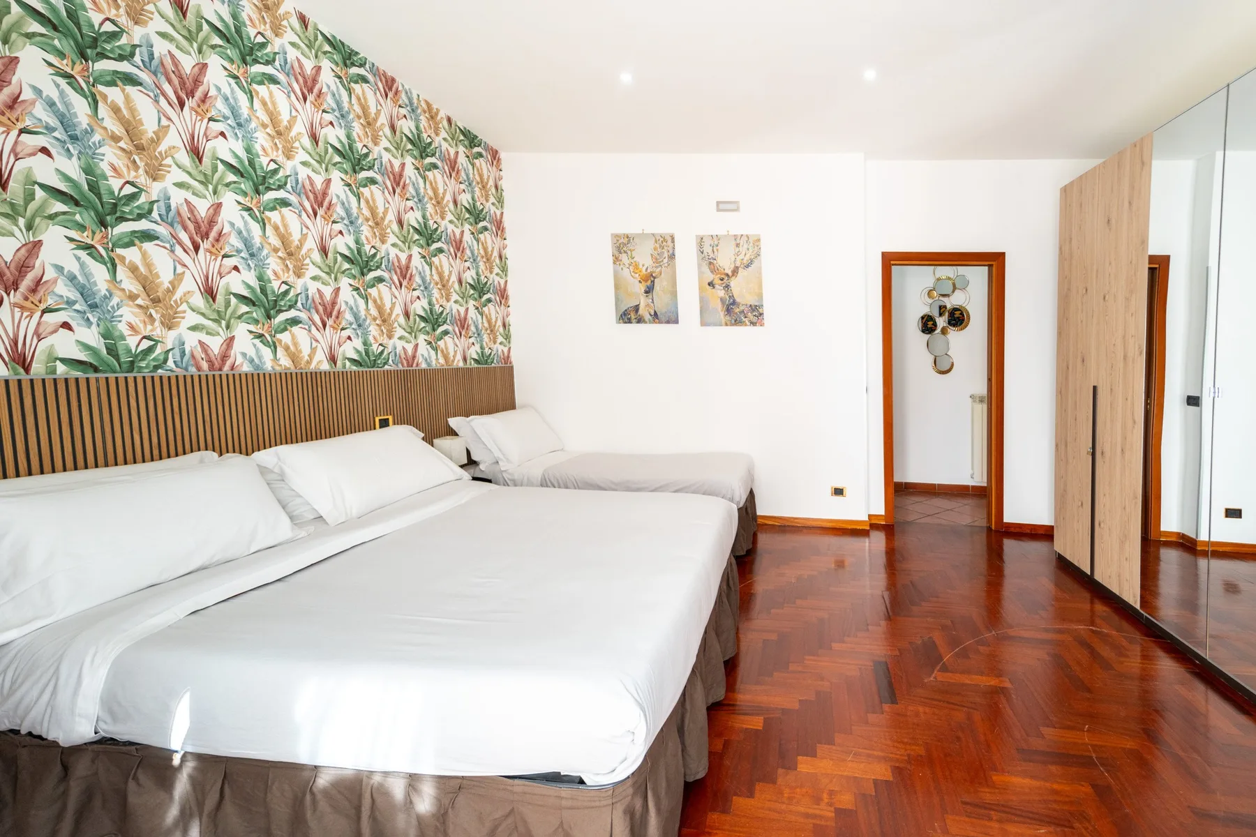 Un dormitorio luminoso con ropa de cama blanca, una pared decorativa botánica y suelo de madera pulida.