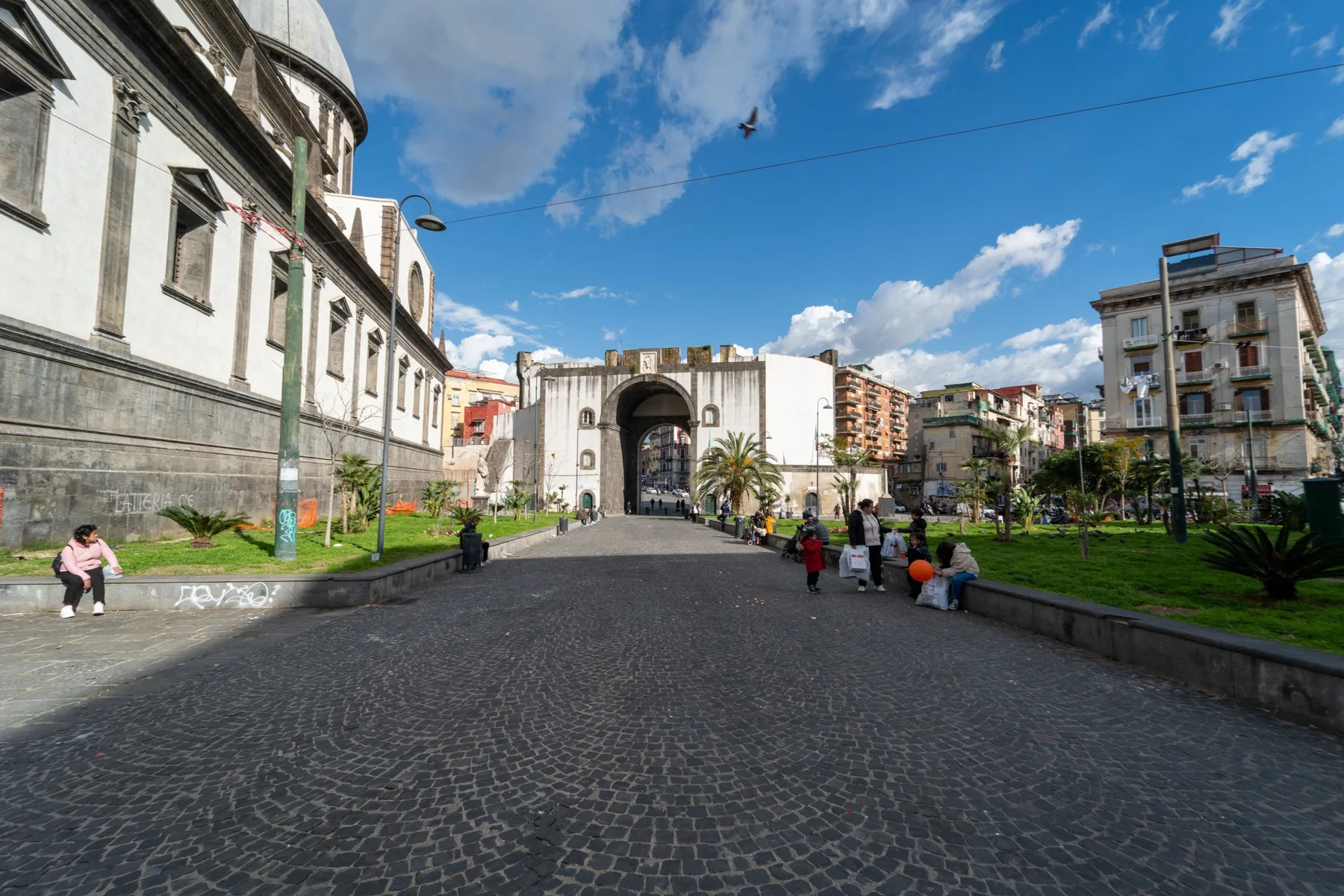 Una plaza empedrada conduce a un arco de piedra, rodeada de edificios y vegetación.