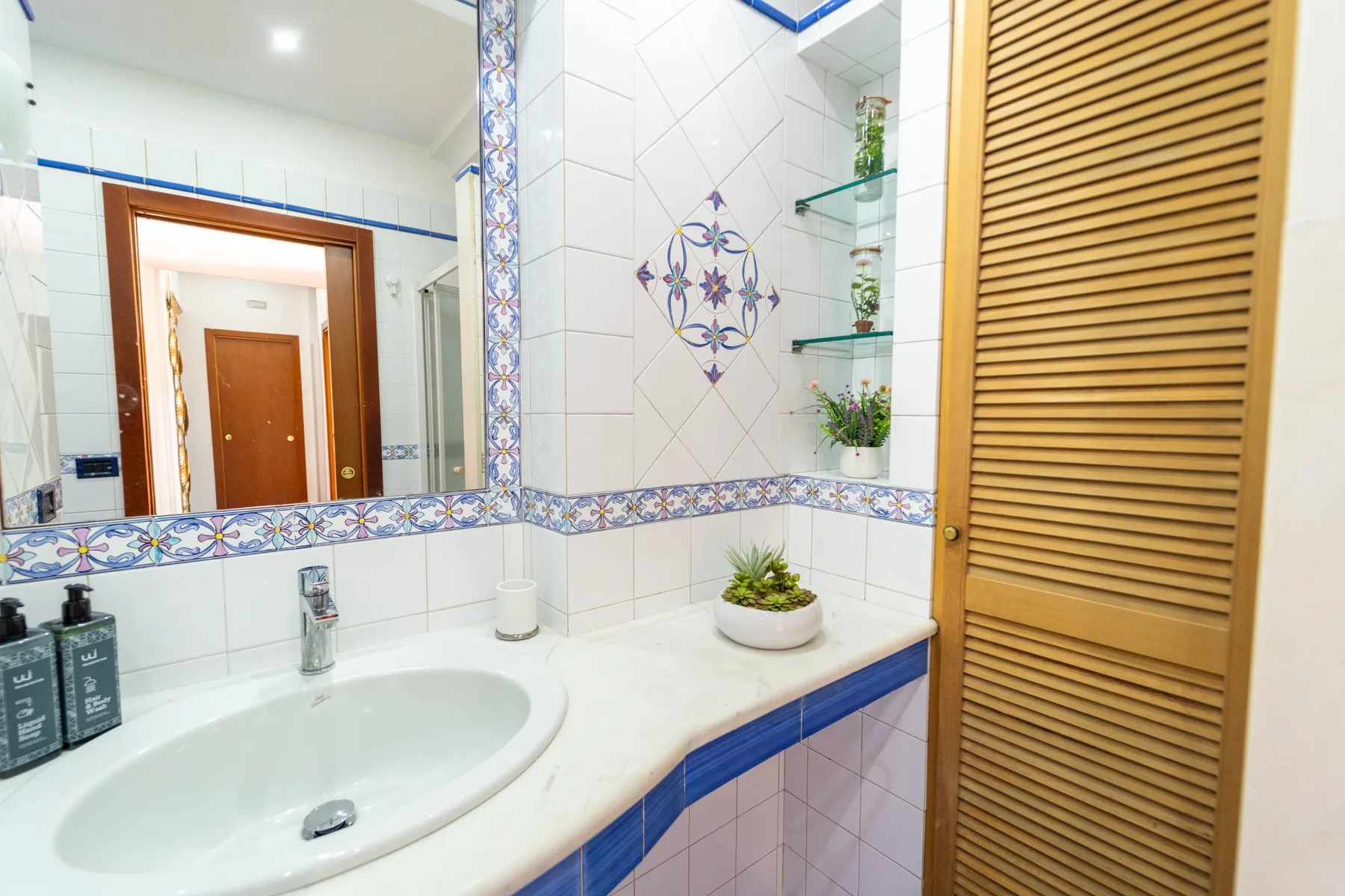 Un baño con azulejos, lavabo, pared con espejo y puerta de lamas de madera.