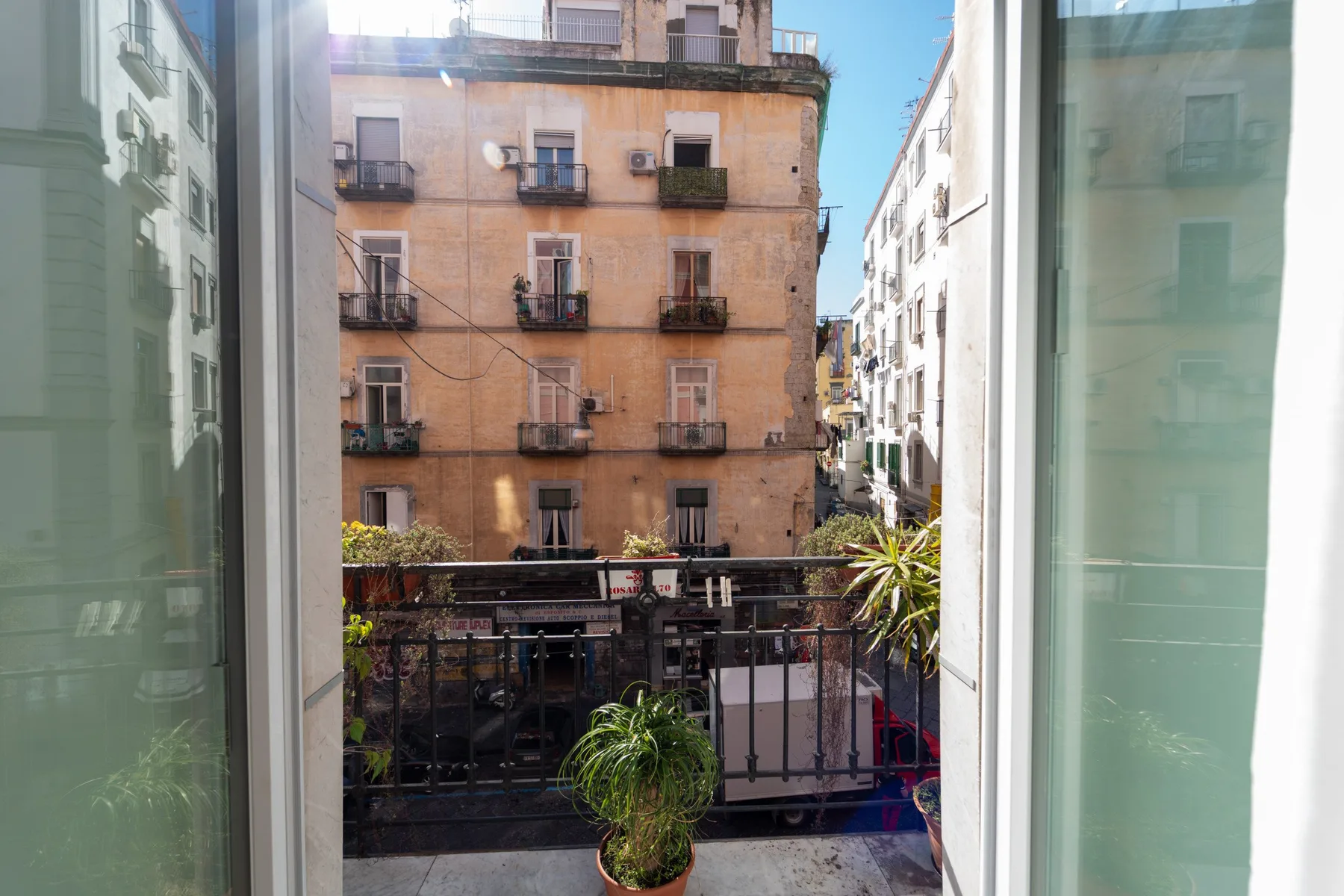 Un balcón con plantas y barandilla da a edificios de apartamentos cercanos.
