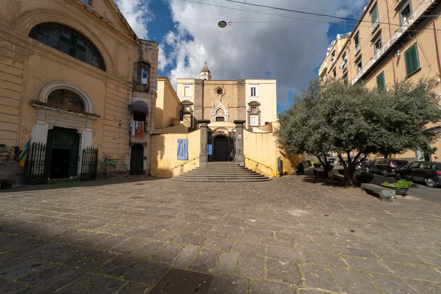 Una plaza pavimentada conduce a un edificio antiguo, con árboles y fachadas alrededor.