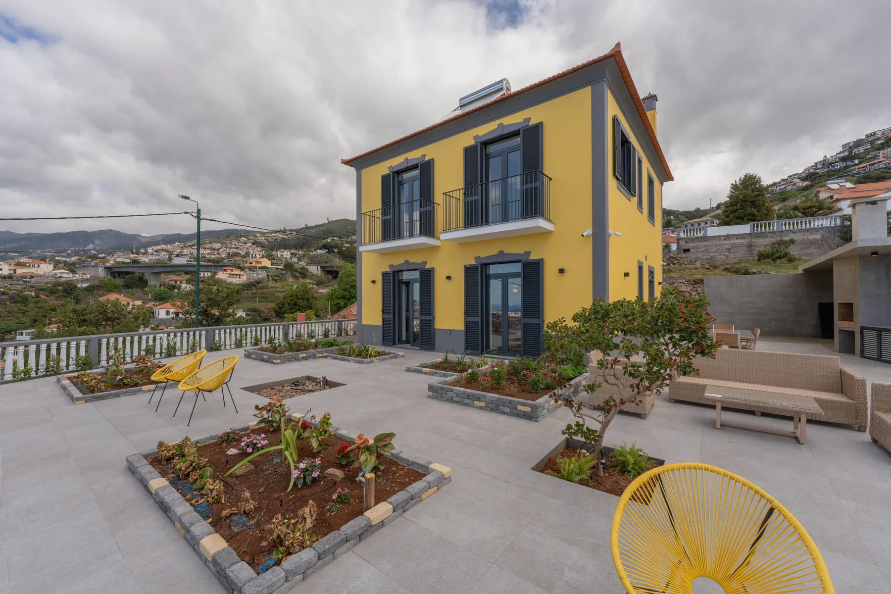 Una casa amarilla está junto a una terraza pavimentada con parterres y asientos al aire libre.