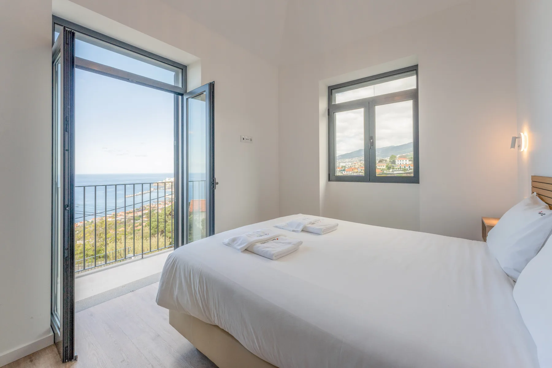 Un dormitorio luminoso con una cama blanca y una puerta abierta al balcón con vistas al mar.