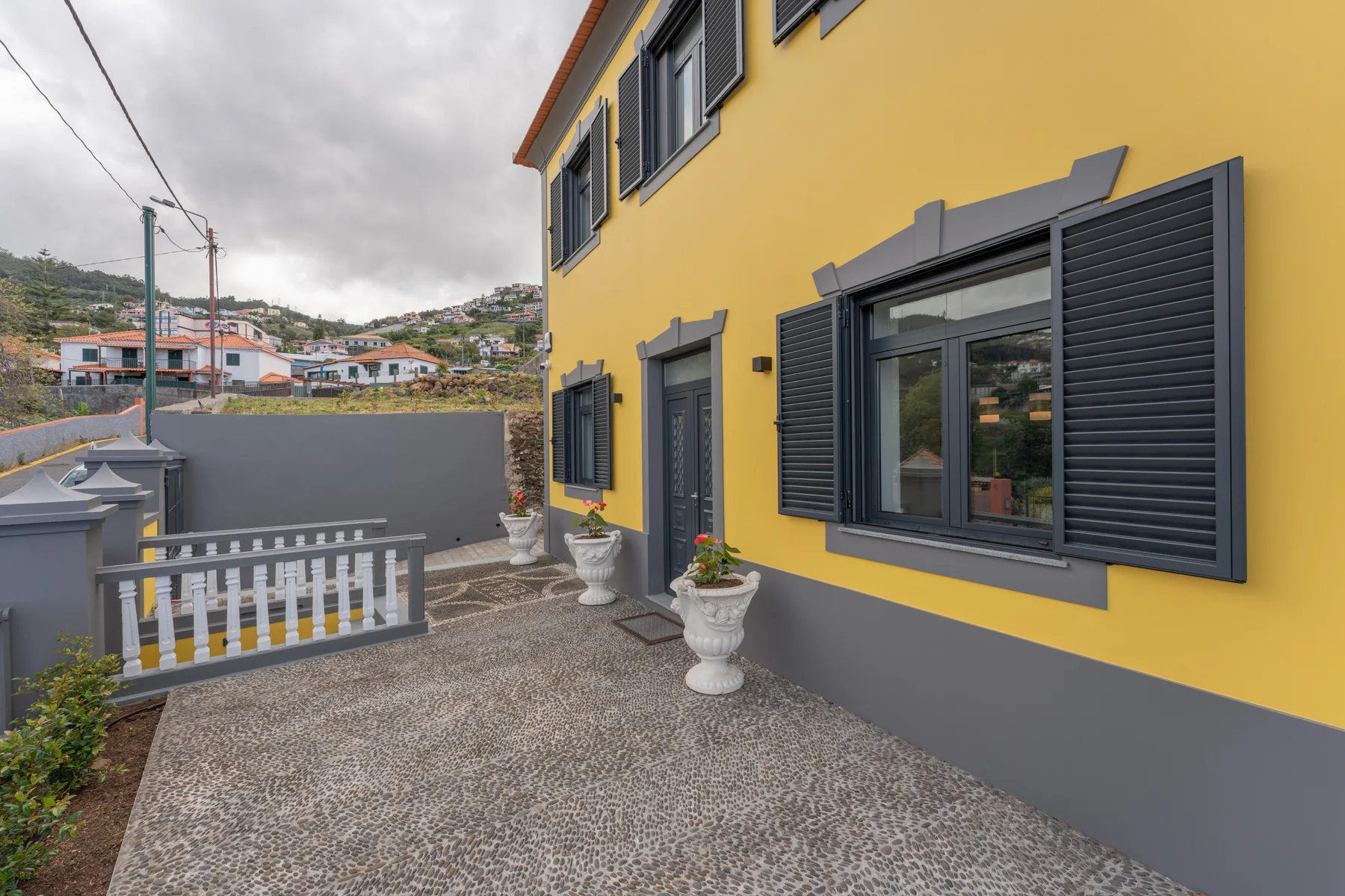 El exterior de una casa amarilla da a una terraza pavimentada con ventanas con contraventanas.