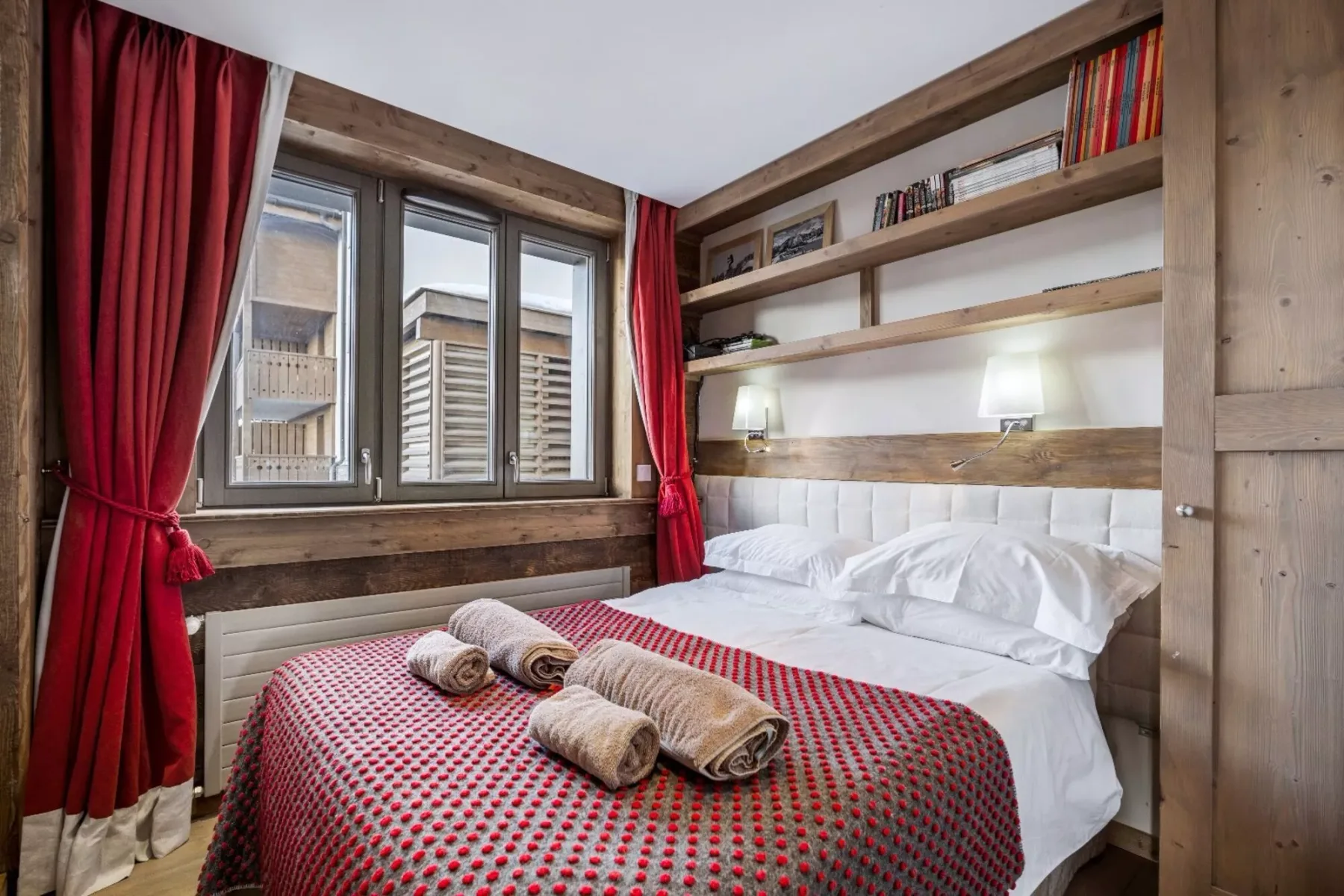A bedroom with a bed, red curtains, and wooden shelves along the wall.